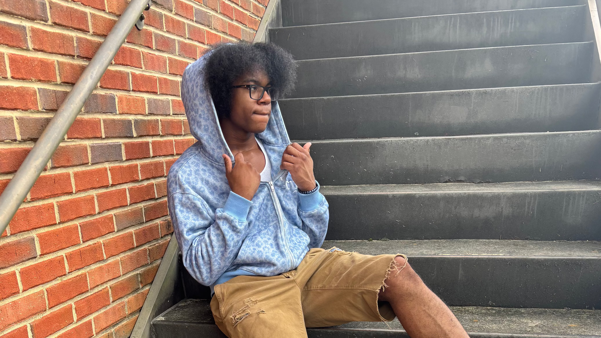 A person sitting on outdoor concrete stairs next to a red brick wall, wearing a light blue hooded sweatshirt, tan shorts, and white sneakers with blue accents.